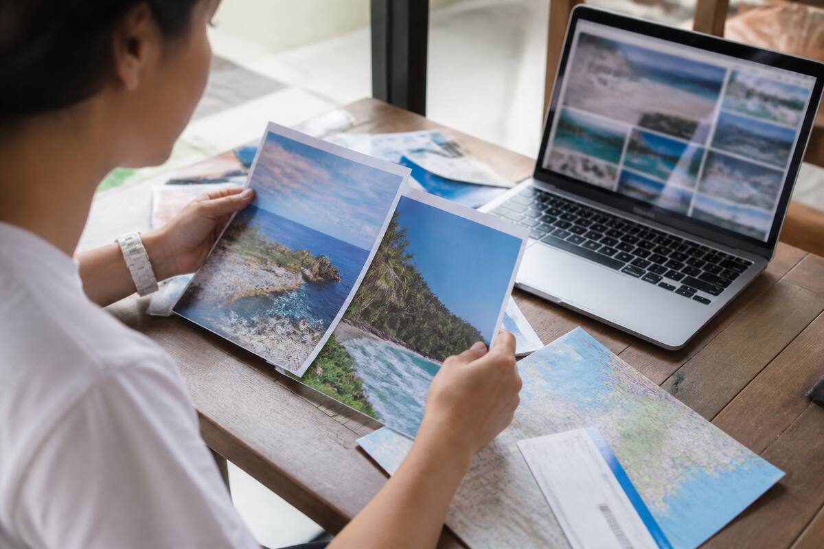 Iemand die vakanties vergelijkt met een laptop en reisbrochures op een houten tafel, bij daglicht